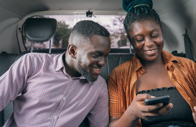 An image of a man and a lady sitting at the backseat of a car. the man is looking at the go-ride app the lady is showing to her